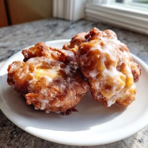 Three golden brown, freshly fried apple fritter pastries drizzled with white glaze sitting on a small white plate.