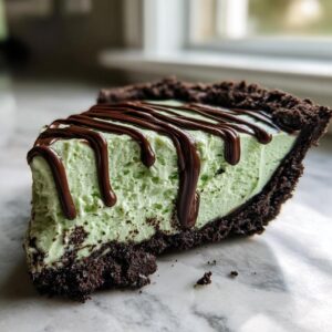 A close-up of a slice of creamy, light green grasshopper pie with a dark chocolate crust and chocolate syrup drizzled on top.
