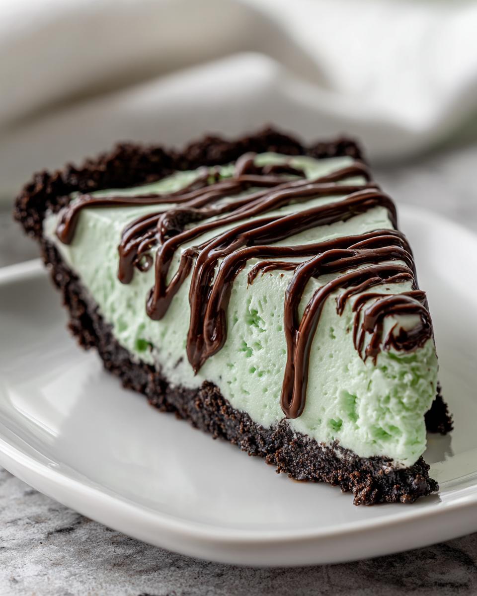 A close-up of a slice of creamy, light green grasshopper pie with a dark chocolate cookie crust and chocolate syrup drizzled on top.