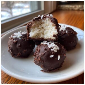 Four rich, chocolate-coated almond joy truffles stacked on a white plate, one cut open showing the shredded coconut filling.