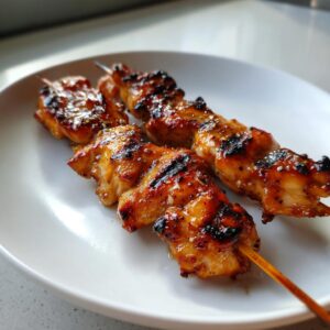 Close-up of two perfectly grilled honey garlic chicken skewers with a shiny glaze and char marks, resting on a white plate.
