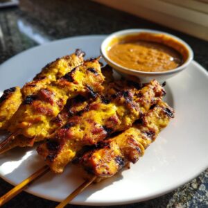 Close-up of perfectly grilled, golden chicken satay skewers served with a side of rich peanut dipping sauce.