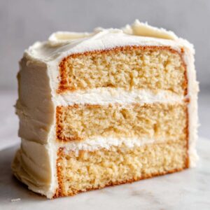 Close-up of a moist slice of three-layer vanilla cake recipe with thick white buttercream frosting.
