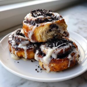 A stack of three gooey oreo cinnamon rolls drizzled with white icing and topped with crushed cookies.