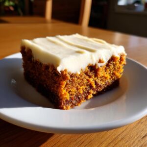 A square slice of moist pumpkin bars with thick cream cheese frosting on a white plate.