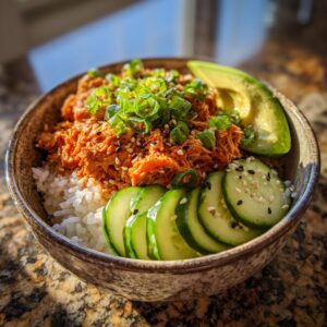 Close-up of a delicious tuna rice bowl topped with spicy tuna mixture, green onions, sesame seeds, sliced cucumber, and avocado.
