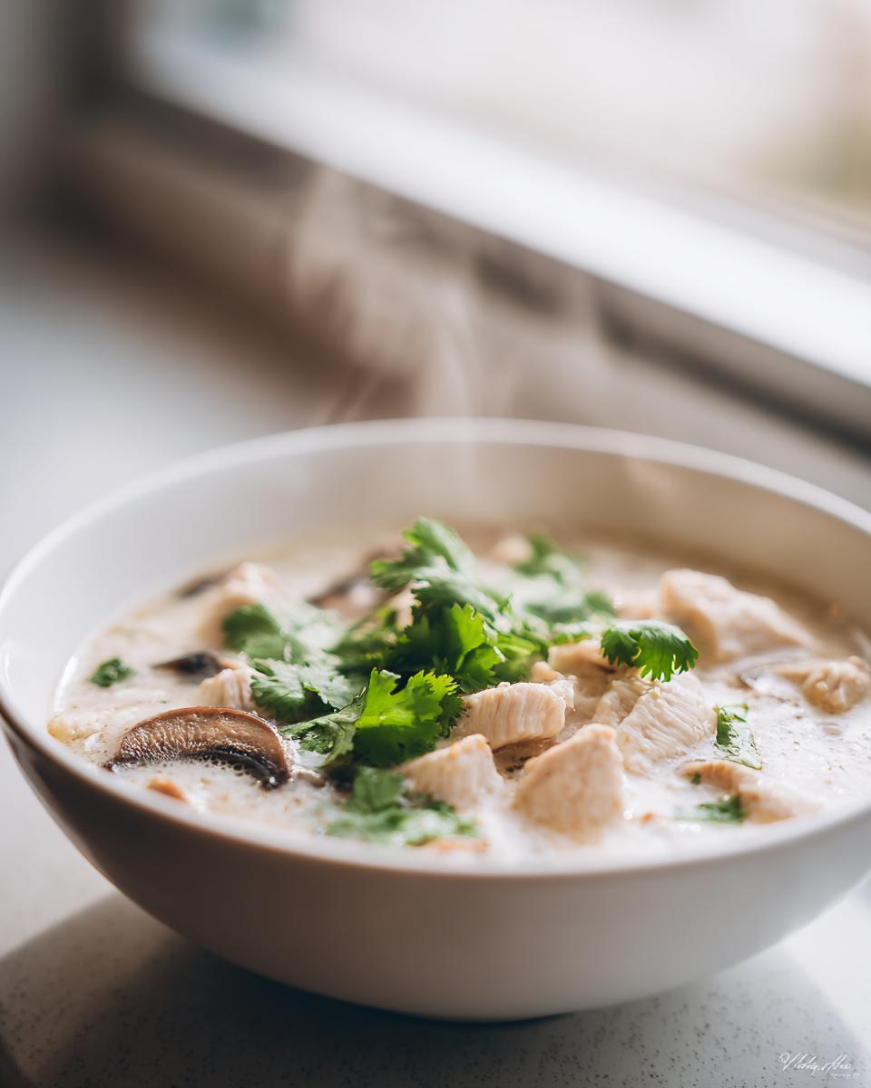 Close-up of a steaming white bowl filled with creamy chicken coconut soup, topped with fresh cilantro and mushrooms.