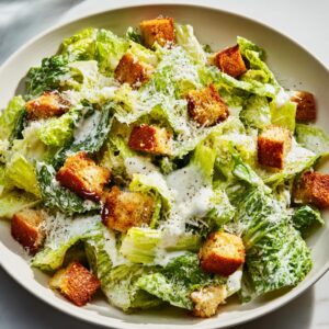 A close-up, overhead view of a bowl filled with fresh caesar salad, tossed in creamy dressing, topped with large croutons and grated Parmesan.