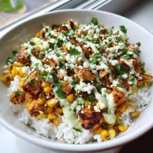A close-up view of a vibrant chicken rice bowl featuring grilled chicken, corn, white rice, crumbled white cheese, and a light green sauce.