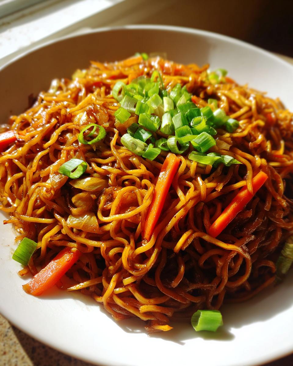 A close-up serving of glossy, dark brown fried noodles topped with bright green chopped scallions and julienned carrots.