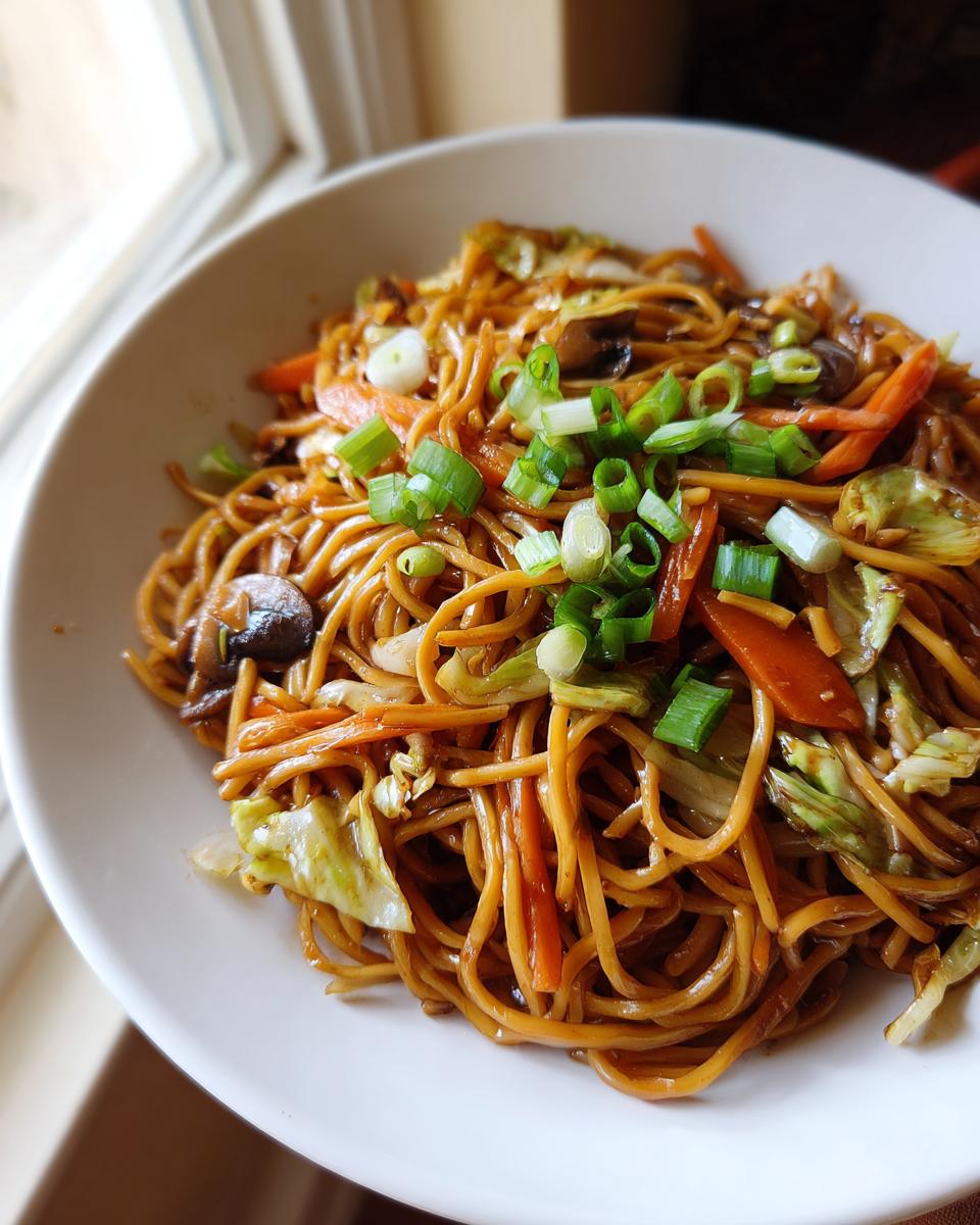 A close-up shot of a white bowl filled with glossy, savory fried noodles mixed with carrots, cabbage, and mushrooms, topped with fresh green onions.