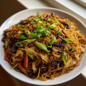 A close-up of a white bowl filled with savory fried noodles mixed with mushrooms, carrots, and cabbage, topped with fresh green onions.