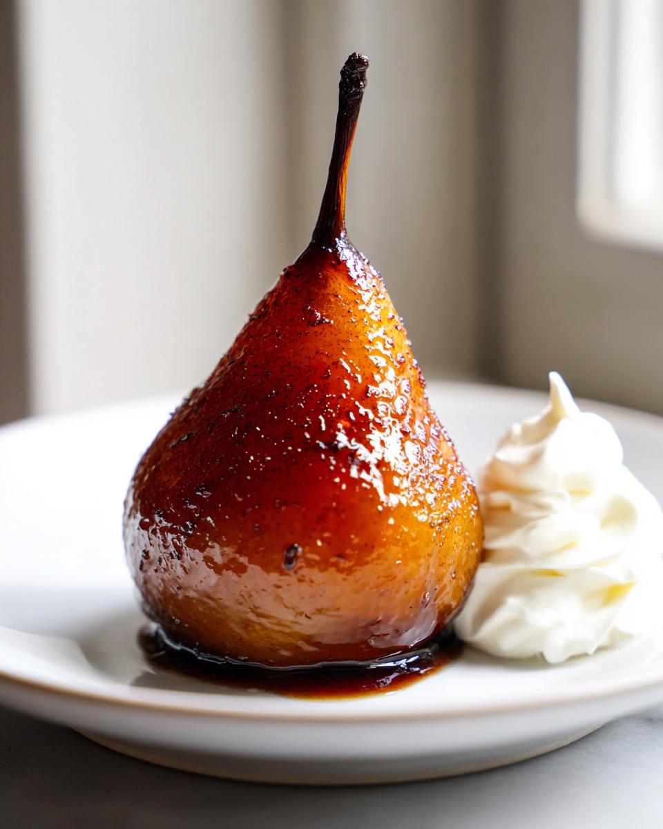 A single, beautifully caramelized poached pear served with a dollop of fresh whipped cream on a white plate.