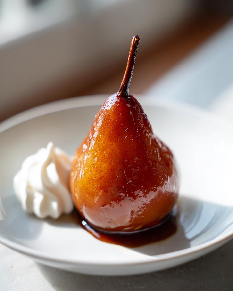 A single, glistening, amber-colored poached pear served on a white plate next to a dollop of whipped cream.