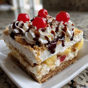 A delicious, layered slice of banana split cake topped with whipped cream, chocolate syrup, and maraschino cherries.