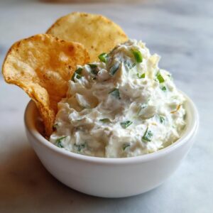 A small white bowl filled with creamy clam dip topped with green onions, served with potato chips.