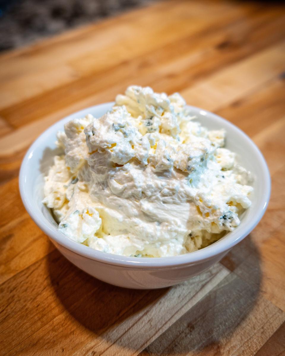 A close-up of chunky, creamy blue cheese dip with visible blue specks in a small white bowl on a wooden surface.