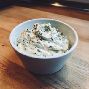 A small white bowl filled with creamy, chunky blue cheese dip mixed with visible green herbs, sitting on a wooden cutting board.