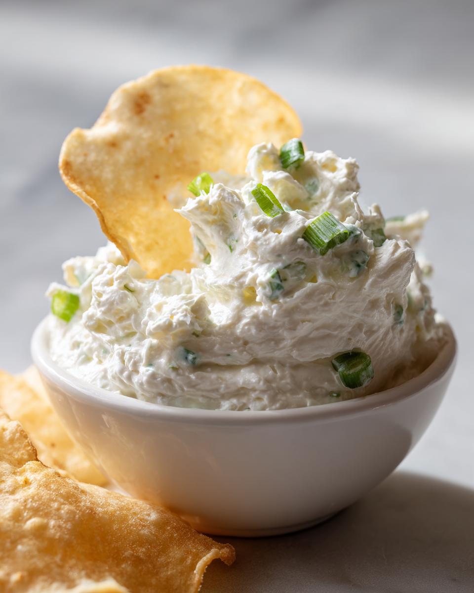 A close-up of creamy clam dip topped with chopped green onions, served in a white bowl with a chip dipped in.