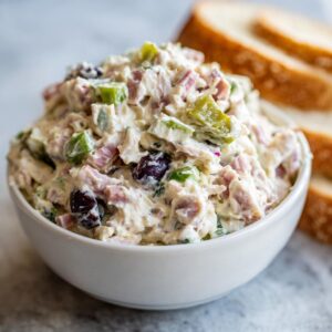 Close-up of a white bowl filled with creamy hoagie dip containing ham, peppers, and olives, with bread slices in the background.