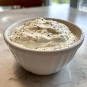 A small white bowl filled with thick, white creamy horseradish sauce, speckled with black pepper and herbs.