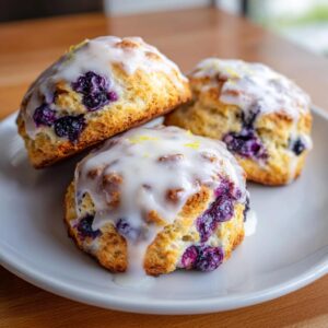 Three freshly baked lemon blueberry scones topped with thick white glaze and lemon zest on a white plate.