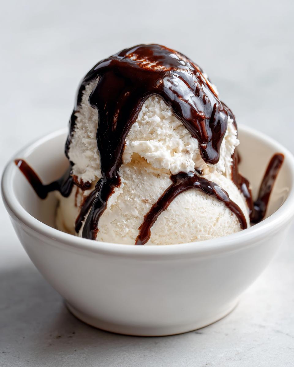 Two scoops of vanilla ice cream in a white bowl generously topped with homemade chocolate syrup.