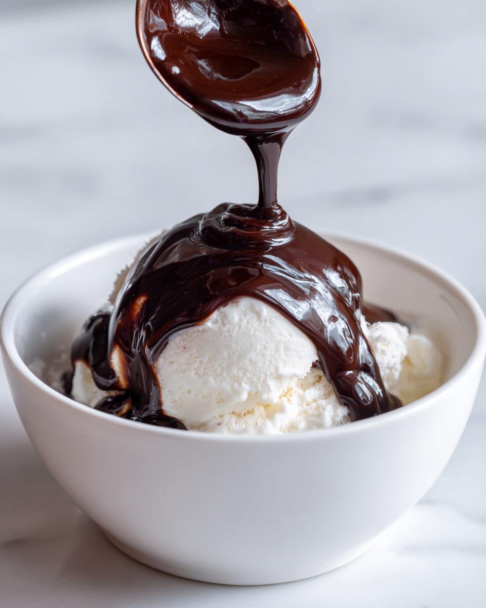 A spoonful of rich, homemade chocolate syrup being poured over scoops of vanilla ice cream in a white bowl.