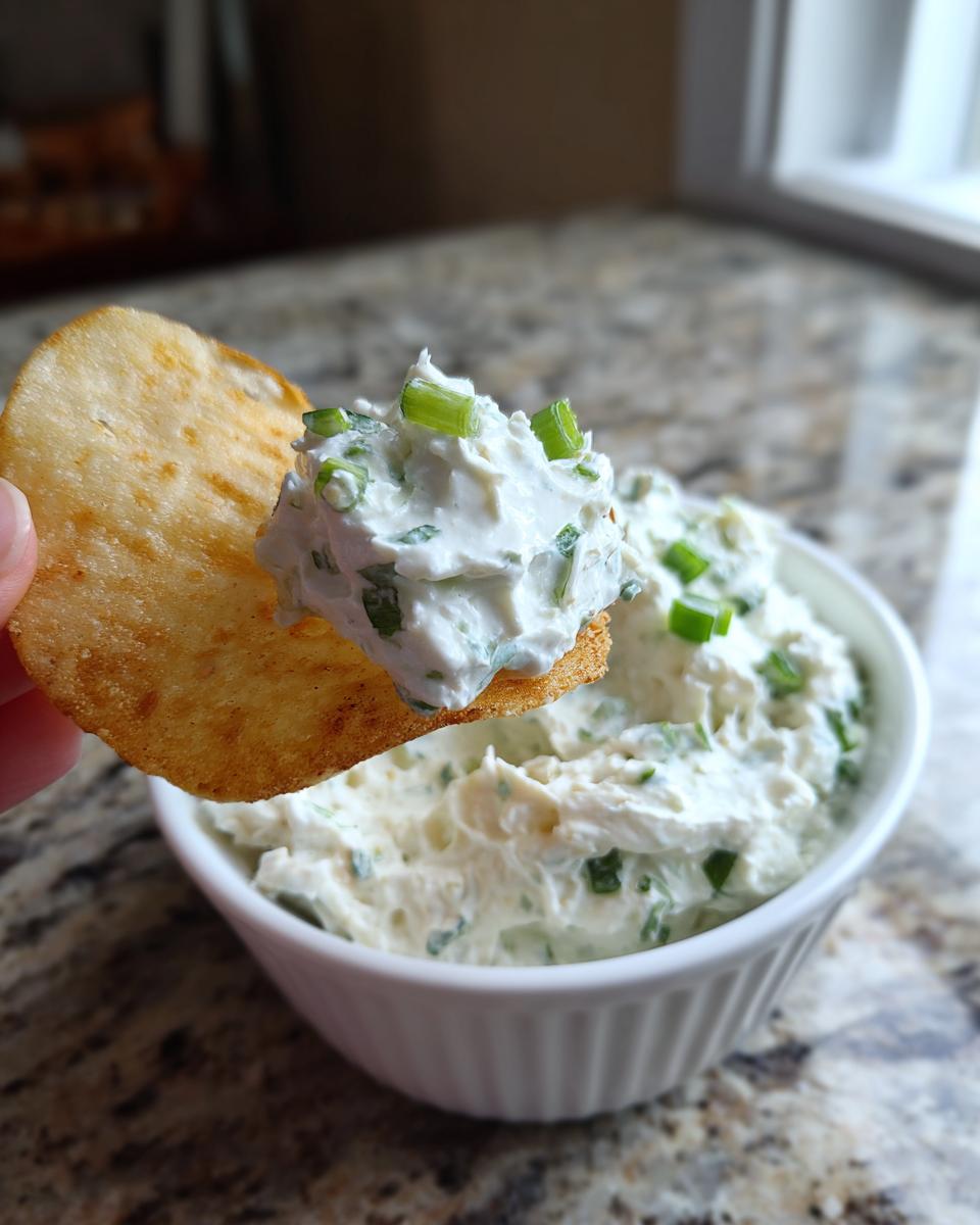 A cracker loaded with creamy clam dip topped with fresh green onions being dipped into a white bowl of clam dip.
