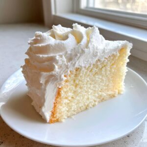 A generous slice of white cake topped high with fluffy, piped vanilla buttercream frosting on a white plate.