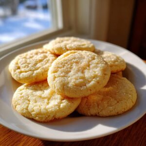 A pile of golden, soft sugar cookies generously coated in sparkling sugar, sitting on a white plate near a window.