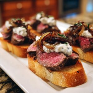 Close-up of three steak crostini bites featuring medium-rare steak, creamy topping, and caramelized onions on toasted bread.