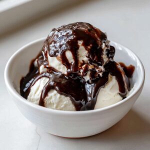 Two scoops of vanilla ice cream generously covered in rich, glossy homemade chocolate syrup in a white bowl.